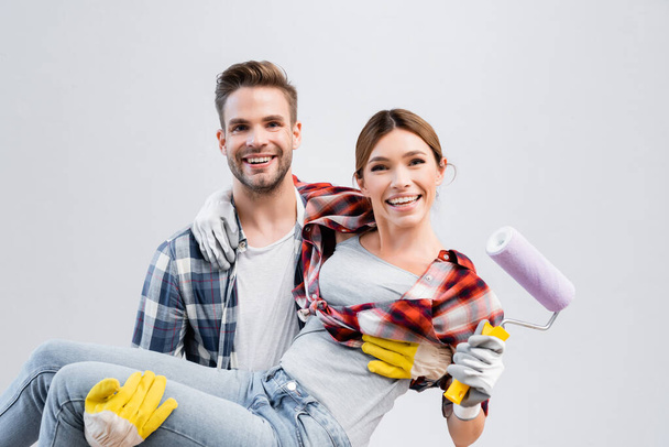 happy young man looking at camera while lifting woman with paint roller  - Photo, image