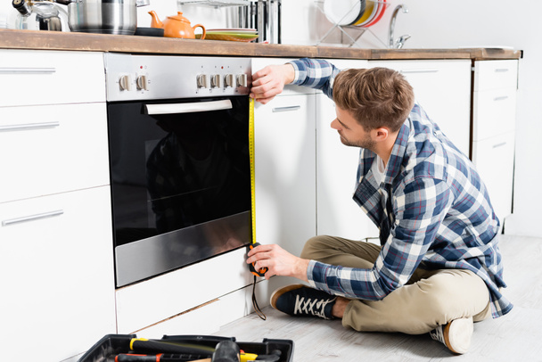 full length of young man with tape measuring oven while sitting near toolbox on floor - Foto, Imagen