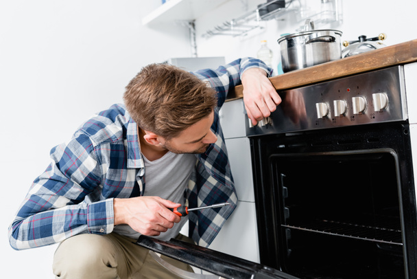young man with screwdriver looking in oven on blurred background in kitchen - Foto, imagen