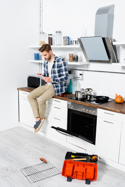 longitud completa de hombre joven usando tableta mientras está sentado en la mesa cerca del horno abierto y caja de herramientas en la cocina - Foto, Imagen