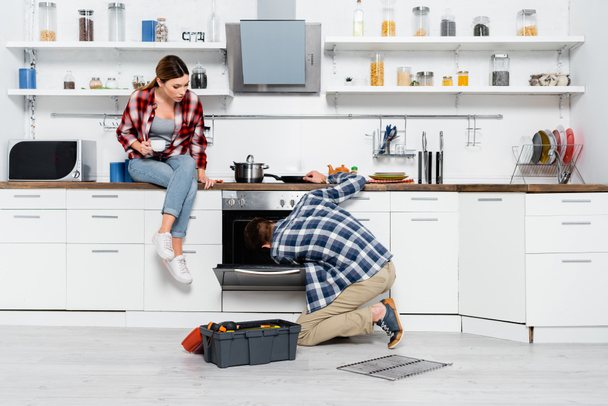 full length of young woman with coffee cup sitting on table near man repairing oven in kitchen - Photo, Image