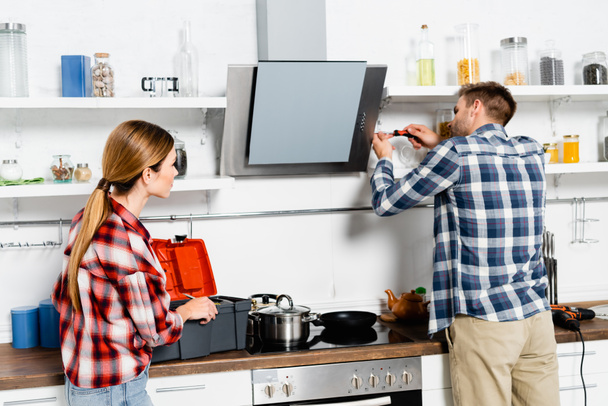 молодая женщина смотрит на мужчину с отверткой ремонт экстрактор вентилятор на кухне - Фото, изображение