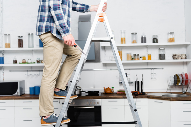 bijgesneden beeld van jongeman met schroevendraaier staande op ladder met wazige keuken op achtergrond - Foto, afbeelding
