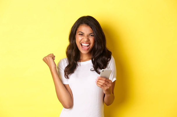 Image of sassy african-american girl in white t-shirt, making fist pump gesture and holding smartphone, celebrating achievement, standing over yellow background - Zdjęcie, obraz