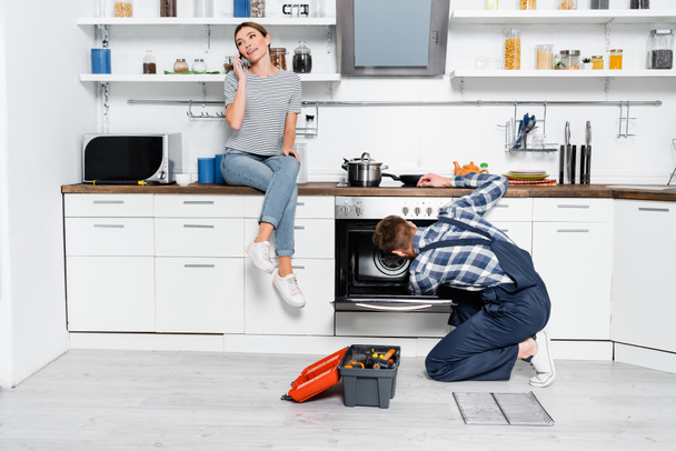 full length of smiling woman talking on smartphone while sitting on table near handyman repairing oven in kitchen - Foto, imagen