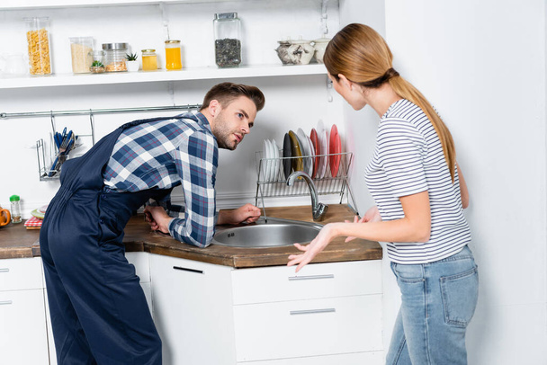 young woman gesturing near handyman leaning on sink in kitchen - Zdjęcie, obraz