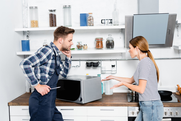 mujer joven señalando con las manos en el microondas cerca de reparador con destornillador en la cocina - Foto, imagen