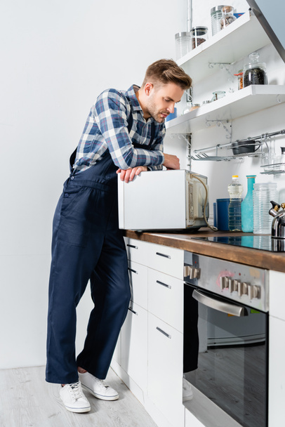 full length of young repairman looking at microwave oven in kitchen - Foto, afbeelding