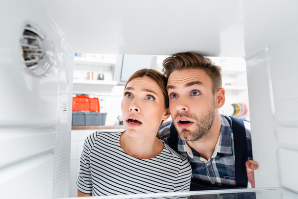 shocked young woman and handyman with open mouths looking at freezer on blurred foreground in kitchen - Photo, Image