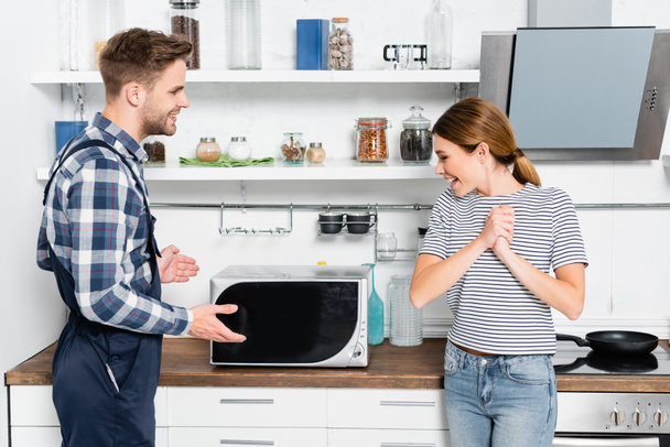 cheerful young woman with clenched hands looking at fixed microwave near handyman in kitchen - Fotografie, Obrázek