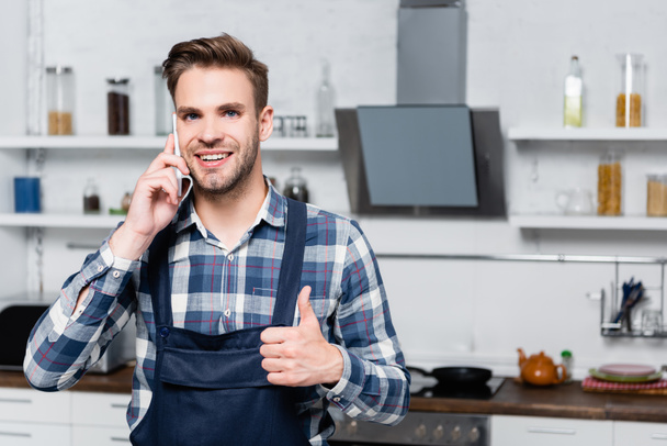 front view of happy man showing thumb up while talking on smartphone with blurred kitchen on background - Foto, Imagem