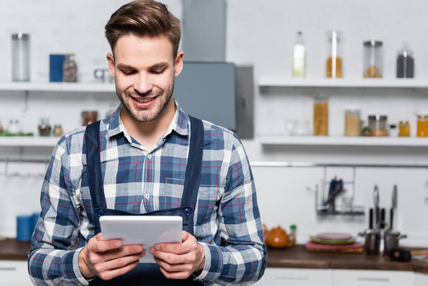 front view of happy handyman holding tablet with blurred kitchen on background - Photo, Image