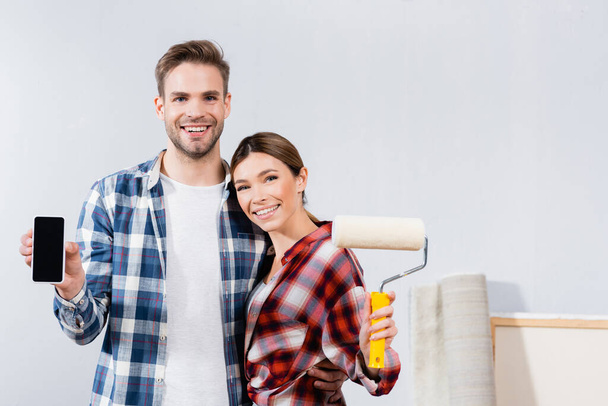 front view of smiling young couple with paint roller and smartphone looking at camera while hugging at home - Foto, afbeelding