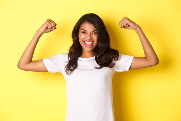 Beeld van zelfverzekerd en vrolijk Afrikaans-Amerikaans meisje, buigende biceps en glimlachend, sterk en assertief, staand over gele achtergrond - Foto, afbeelding