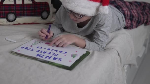 Un niño con una gorra de Papá Noel escribe en papel su deseo de Año Nuevo - Metraje, vídeo