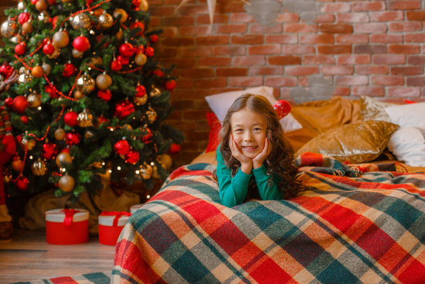 little Asian girl in pajamas in the bedroom lying on the bed near the Christmas tree Christmas, new year - Photo, Image