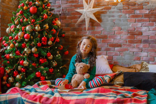 little Asian girl in pajamas in the bedroom lying on the bed near the Christmas tree reading a book with a Teddy bear Christmas, new year - Фото, изображение