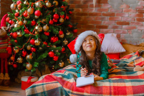 bambina asiatica in pigiama vicino all'albero di Natale scrive una lettera a Babbo Natale, nuovo anno - Foto, immagini