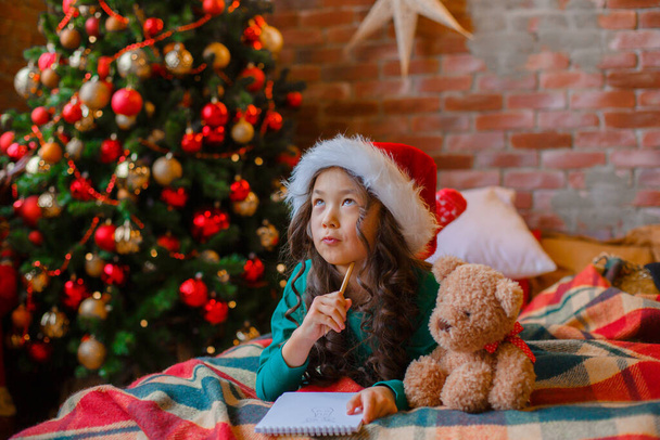 pieni aasialainen tyttö pyjamassa lähellä joulukuusi kirjoittaa kirjeen joulupukille joulu, uusi vuosi - Valokuva, kuva