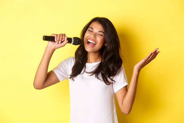 Kaygısız Afrikalı Amerikalı bir kızın mikrofonda şarkı söylemesi mutlu bir şekilde gülümsemesi, sarı arka planda gösteri yapması. - Fotoğraf, Görsel