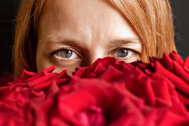 Foto de cerca de una mujer joven con el pelo rojo disfrutando de un gran ramo de rosas rojas. Media cara está escondida por las flores - Foto, Imagen