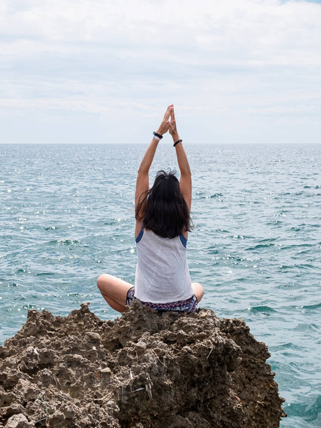 Вид сзади на молодую женщину с длинными волосами, практикующую йогу на скалах в море - Фото, изображение