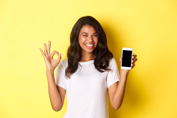 Image of beautiful african-american girl, winking and smiling, showing smartphone screen and okay sign in approval, standing over yellow background - Photo, Image