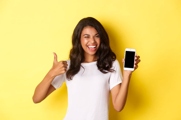 Image of beautiful african-american girl, winking and smiling, showing smartphone screen and thumbs-up in approval, standing over yellow background - Fotografie, Obrázek