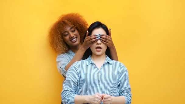 Giovane donna nera afroamericana che copre gli occhi della sua amica asiatica e la sorprende. Espressione facciale eccitata - Foto, immagini