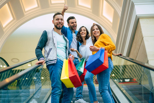 Vier stylische lächelnde aufgeregte schöne moderne Freunde in lässiger Kleidung mit Papiertüten amüsieren sich, während sie beim Einkaufen auf der Rolltreppe im Einkaufszentrum stehen. - Foto, Bild