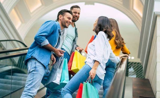 Four stylish smiling excited beautiful modern friends in casual clothes with paper bags are having fun while standing on the escalator in the mall during shopping. - Фото, зображення