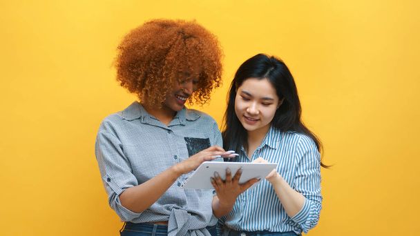 Young multiracial friends browsing social media online with excited face expressions. Afican american and asian women isolated on yellow background - Foto, Imagen