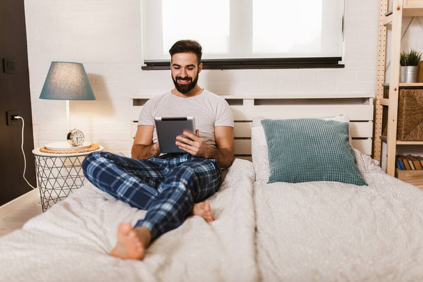 Hezký mladý muž v pyžamu pomocí digitální tablet, zatímco ležel v posteli v dopoledních hodinách - Fotografie, Obrázek
