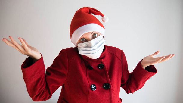 Zweifelnde Frau mit medizinischer Gesichtsmaske zuckt mit den Schultern, sieht unsicher aus, trägt roten Mantel und Weihnachtsmütze. Coronavirus-Pandemie und Weihnachtskonzept. - Foto, Bild