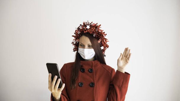 Egy piros kabátos nő orvosi maszkkal és ünnepi szalaggal a fején, telefonnal a kezében és online videóhívással az ünnepek alatt. Közelíts rá a családodra és a barátaidra az influenzaszezon vagy a koronavírus alatt. - Fotó, kép