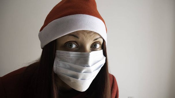 Porträt einer Frau in rotem Mantel und Weihnachtsmann-Weihnachtsmütze mit medizinischer Maske.  - Foto, Bild