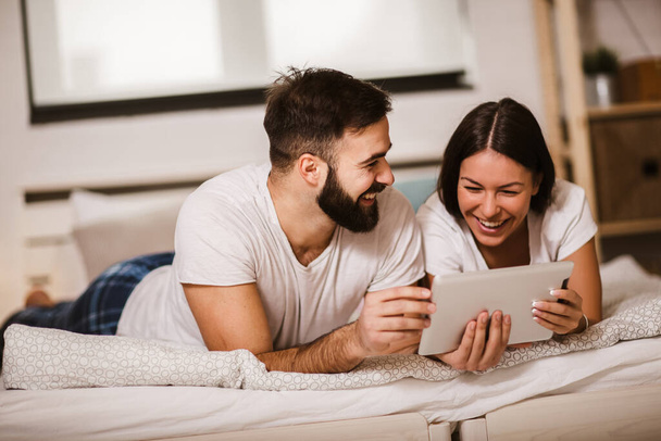 Sonriente pareja joven relajada usando tableta digital en la cama en casa - Foto, imagen