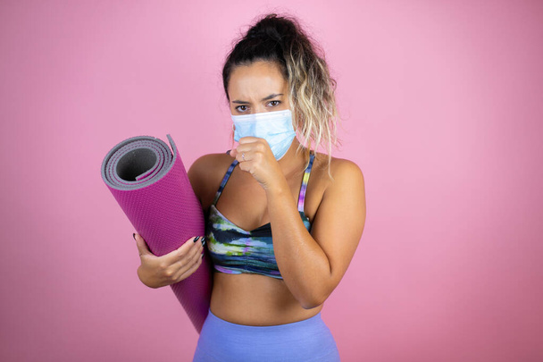 Young beautiful woman wearing sportswear and holding a splinter over isolated pink background with her hand to her mouth because she's coughing with mask - Photo, image