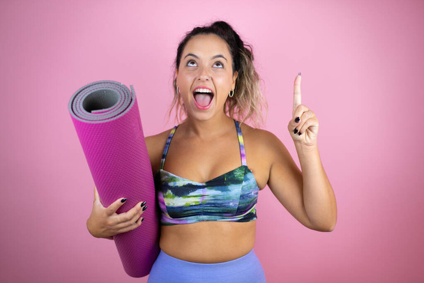 Young beautiful woman wearing sportswear and holding a splinter over isolated pink background amazed and surprised looking up and pointing up with fingers and raised arms - Fotó, kép
