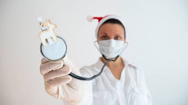 Eine Kinderärztin mit Neujahrsmütze und medizinischer Maske hält ein Stethoskop mit einem Rentier in der Hand. Arztbesuch in den Ferien. - Foto, Bild