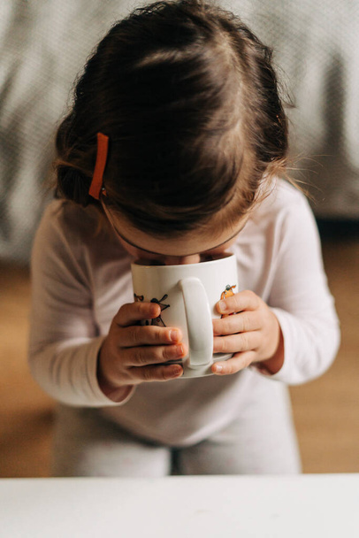 Caucasian toddler girl drinking cocoa from mug at home. Christmas bokeh lights - Foto, imagen