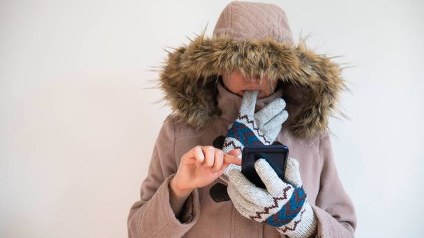 Femme congelée vêtue d'un manteau chaud avec une capuche sur la tête utilise un smartphone. - Photo, image