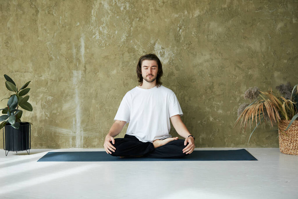 giovane in posa meditazione a casa, bel maschio seduto in loto su tappetino yoga con piante, copiare lo spazio - Foto, immagini