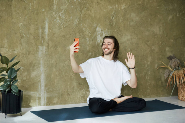 nauczyciel medytacji na telefon wideo, przystojny mężczyzna siedzi w jogi asana i ma lekcje wideo przez telefon - Zdjęcie, obraz