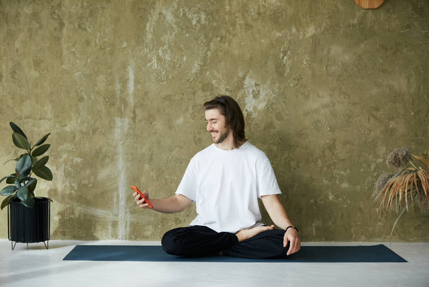 mężczyzna siedzi w pozycji do jogi na macie za pomocą smartfona, przystojny mężczyzna uśmiecha się i trzyma telefon podczas jogi i medytacji - Zdjęcie, obraz