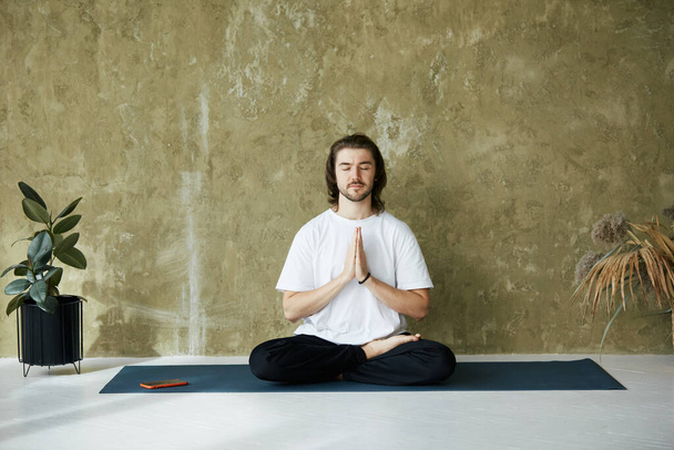 handsome calm man sitting in lotus asana and namaste mudra, male in white shirt with calm relaxed emotion holding arms in namaste - Foto, afbeelding