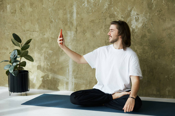 lovely man in yoga pose on mat using phone,remote practice of yoga or meditation online through phone, copy space - Photo, Image