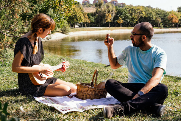 Пара сидить на скатертині насолоджуючись пікніком, граючи в укулеле і фотографуючи
 - Фото, зображення