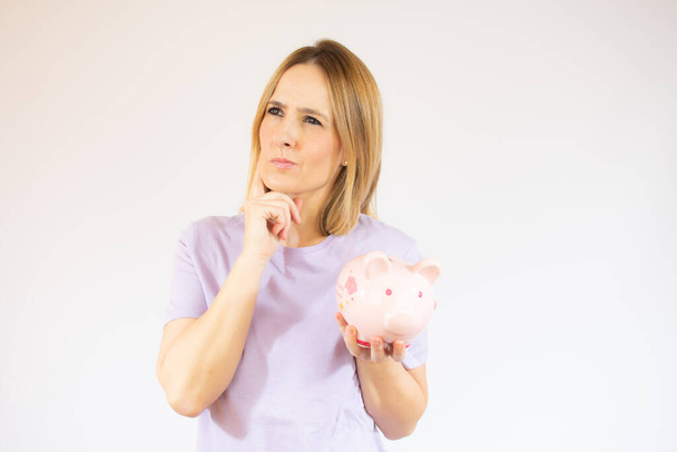 Portrait of a pensive casual woman looking away at copy space holding a piggy bank isolated over white background - Fotoğraf, Görsel