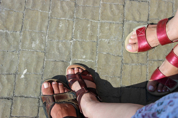 Παπούτσια ποδιών ανδρών και γυναικών σε σανδάλια, σε πέτρινο πεζοδρόμιο στο φως του ήλιου - Φωτογραφία, εικόνα